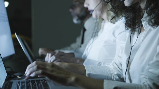 Call center operators using laptops. Side view of male and female colleagues in headsets talking and using computers in dark office, slider shot. Customer support concept
