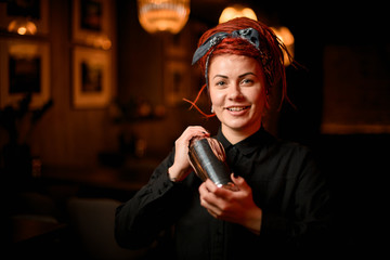 Cute smiling redhead bartender girl with dreadlocks standing in the bar with a steel shaker