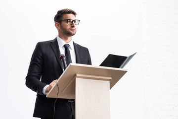 businessman in suit standing at podium tribune and looking away during conference isolated on white