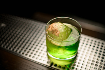 Alcoholic green transparent cocktail in the glass with the one big ice cube and spices on it