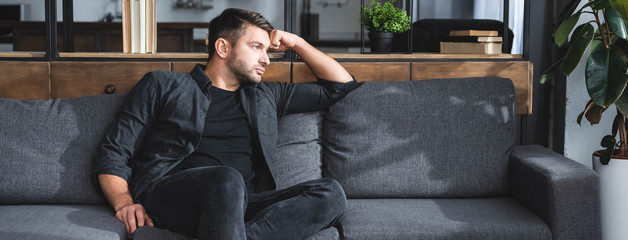 panoramic shot of handsome and pensive man sitting on sofa in apartment