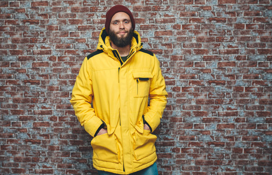 City portrait of handsome hipster guy with beard wearing a blank winter yellow jacket and red hat standing on a brick wall background. Empty space for your logo or design. Mockup for print.