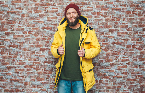 City portrait of handsome hipster guy with beard wearing a blank winter yellow jacket and red hat standing on a brick wall background. Empty space for your logo or design. Mockup for print.
