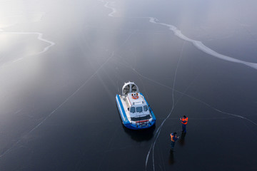 The river is covered with thin ice. On the icy surface is a hovercraft next to the boat are coast guard rescuers. The view from the top. - obrazy, fototapety, plakaty