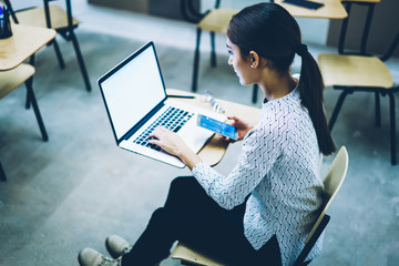 Back view of female student synchronizing telephone files with laptop computer sitting at desk,clever young woman searching training webinar on netbook with blank screen for getting information