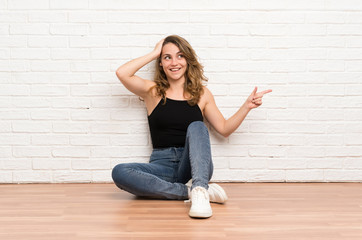 Young blonde woman sitting on the floor surprised and pointing finger to the side