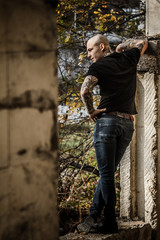 Bald brutal young man with tattoos stuffed on his arms and neck against the background of an abandoned destroyed building. Attractive tattooed stern guy. Concept of a bully and a dangerous guy.