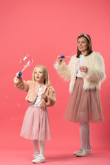daughter and smiling mother blowing soap bubbles on pink background