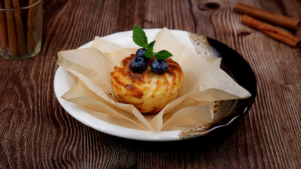 Blueberry muffin with fresh berries and mint leaves on top in waxed paper on a plate on a wooden table next to cinnamon sticks
