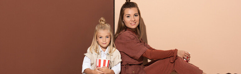 panoramic shot of daughter with popcorn and smiling mother looking at camera on beige and brown background