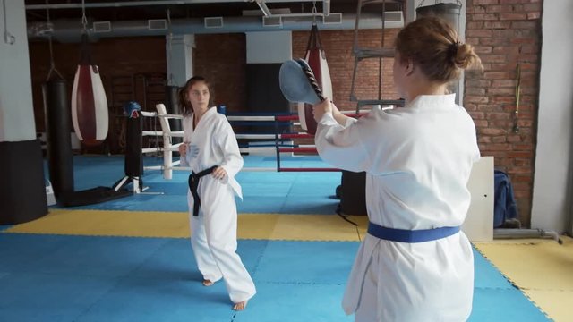 Pan shot of young barefoot attractive woman wearing white kimono with black belt practicing kicks with the help of female fighter holding special equipment in her hands