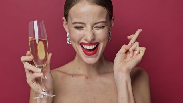 Pretty young woman with red lipstick and earrings is making a wish while holding a glass of champagne isolated over red background