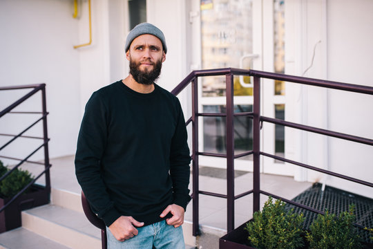 City portrait of handsome hipster guy with beard wearing black blank hoody or sweatshirt and hat with space for your logo or design. Mockup for print