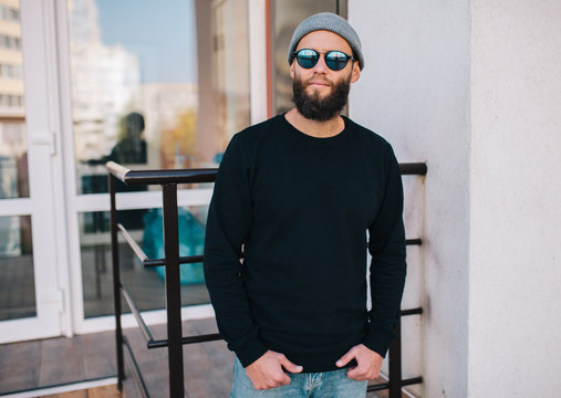 City portrait of handsome hipster man with beard wearing black blank hoody or sweatshirt and hat with space for your logo or design. Mockup for print