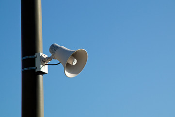 A white loud speaker or siren is mounted on a pole against a clear blue sky with copy space.
