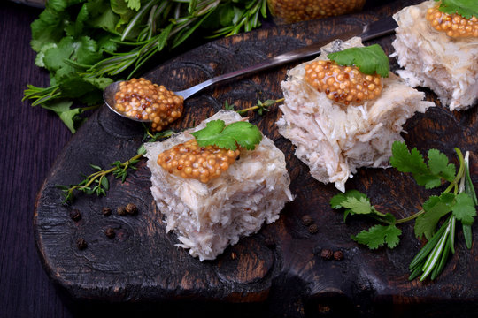 Holodets chicken jelly cut into cubes topped with mustard and cilantro on a dark wooden board surrounded by herbs and pepper