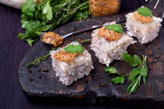 Holodets chicken jelly cut into cubes topped with mustard and cilantro on a dark wooden board surrounded by herbs and pepper