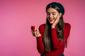 Pretty european young woman in red holding small jewelry box with proposal diamond ring on pink wall background. Girl smiling, she is happy to get present, proposition for marriage.