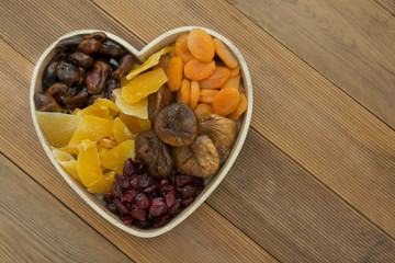 Dried fruits mix, in wooden heart shape box isolated on wooden background. Top view of various dried fruits figs, apricots, mango, cranberries. Copy space.