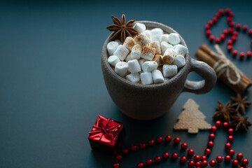 Christmas background. Marshmallow cocoa in clay bowl sprinkled with cinnamon, anise, cinnamon sticks with hemp rope and red christmas modern decoration as gift, beads and fir tree at blue background.