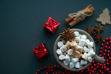 Fototapeta na wymiar Christmas background. Marshmallow cocoa in clay bowl sprinkled with cinnamon, anise, cinnamon sticks with hemp rope and red christmas modern decoration as gift, beads and fir tree at blue background.