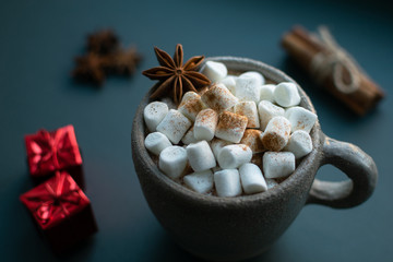 Hot chocolate with marshmallow sprinkled with cinnamon, with anise star, cinnamon sticks tied up with hemp rope and red christmas decorative gifts at blue background. Winter beverage.