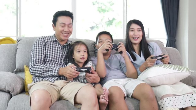 Asian Family playing videogame holding joysticks sitting on sofa at home enjoying game. Modern lifestyle, happy youth and relationship concept. Happuy family relaxing on sofa together at home.