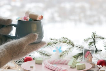 Obraz na płótnie Canvas A cup with a drink and marshmallows in hands in gloves on the background of Christmas decor, illumination on a wooden windowsill, the concept of home comfort, winter holidays