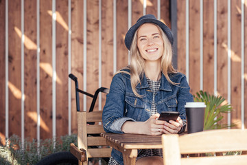 Stylish young student girl in denim clothes chatting in social networks using a smartphone while sitting outdoor at wooden table of an open cafe in the warm summer time