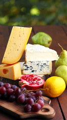 Camembert, brie, hard cheese and grapes on wooden stand. Pieces of different cheeses, pear and lemon on blurred background. Dorblu, soft cheese and pomegranate on boards. Widescreen