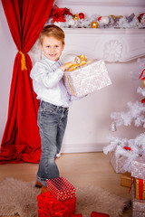 Obraz na płótnie Canvas beautiful little boy in a light shirt and jeans smiles and holds in his hands a big gift with golden bows, around presents, New Year tree, red-white festive Christmas background.