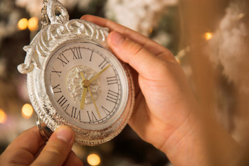 Close up, macro. Children's hands are holding a vintage white clock. Against the background of a Christmas tree decorated with garlands. Copy space.