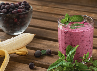 Blackcurrant smoothie with banana and mint in a glass cup. Smoothie is decorated with mint leaves, a glass is on a brown wooden table. Nearby ingredients for smoothies. Close-up.