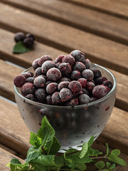Glass bowl with blackcurrant berries in a frost on a dark wooden table. Nearby are mint leaves and blackcurrant berries, ingredients for smoothies. Close-up. Vertical frame