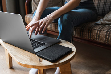 Woman is sitting on a sofa and using a laptop at wooden table. Study and work online, freelance. Self employed girl is working with her notebook sitting on a couch with a phone and ereader on table.