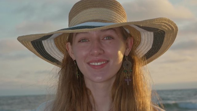 Young woman with a hat smiling at the camera during sunset by the ocean