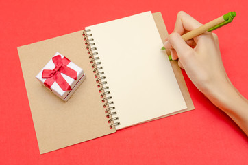 hand of a girl who is going to draw on a blank sheet in a notebook, next to a gift box on a red...