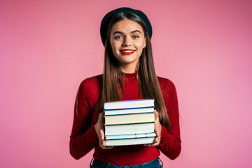 Pretty student on pink background in studio holds stack of university books from library. Girl in...