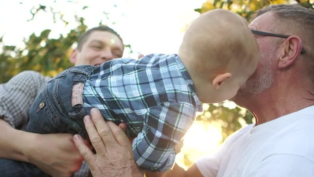 Grandfather, son and grandson have fun in the apple orchard park. A young man holds his little son in his arms, grandfather holds out his hands to the baby