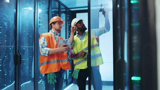 Diverse team of IT technicians fixing bugs and errors in server cabinet, using digital tablet and wearing high vis safety vests. Professional team. Data center.