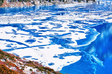 Last colors of the passing autumn: a multi-colored shrub on the slope contrasts with blue water and bizarre forms of ice on a frozen mountain lake; beauty and variety of shapes in nature concept