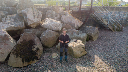 An aerial view of a happy smiling midage male drone pilot with hat along a stony beach and massive rock boulder in the background