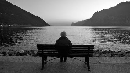 A lonely old man sits on a wooden bench at dusk after sunset, looking at the lake and the light on...