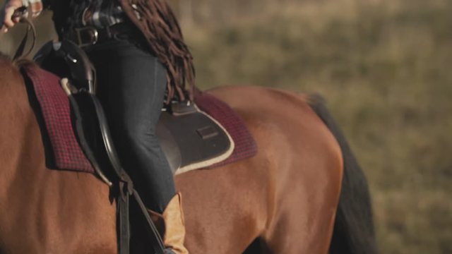 Close up of young cowgirl in saddle at brown horse outdoors. Beautiful woman riding horse at sunset field.