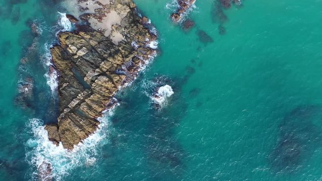 Aerial view of crashing waves on rocks and beach. Drone view of sea waves and beautiful rocky coast. Background texture of a sandy shore and blue turquoise water. Flying upwards.