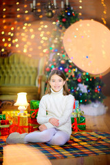 Fototapeta na wymiar A funny girl sits on the floor next to gifts on the background of bright holiday lights. Christmas and New Year.