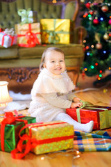 Fototapeta na wymiar A cute girl in a white dress is sitting on the floor next to gifts and smiling cheerfully. Christmas and New Year
