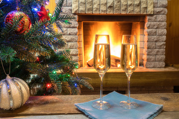 Christmas eve. Two glasses of champagne wine near cozy fireplace and before Christmas tree decorated toys and christmas lights in country house.
