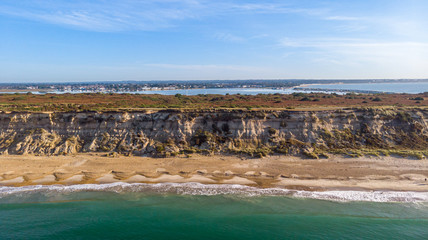 An aerial view of a majestic seacoast hill with huge cliff, sandy beach, groyne (breakwater) and crystal blue water under a beautiful blue sky and some white clouds