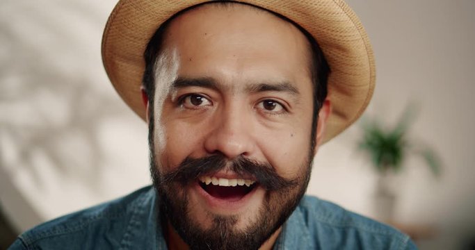 Close up of super surprised emotional Hispanic male with mustache and beard indoor in hat, cheerful brunette guy smiling of unexpected news showing expressive facial expression dressed in jeans shirt